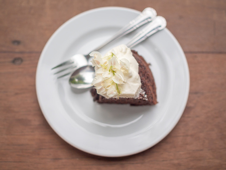 A piece of wild organic chocolate honey cake with beautiful vintage flowers topping and whipping cream on top in white ceramic plate with sillver spoon and fork on wooden table background top view.の写真素材