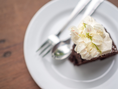 A piece of wild organic chocolate honey cake with beautiful vintage flowers topping and whipping cream on top in white ceramic plate with sillver spoon and fork on wooden table background top view with copy space.の写真素材
