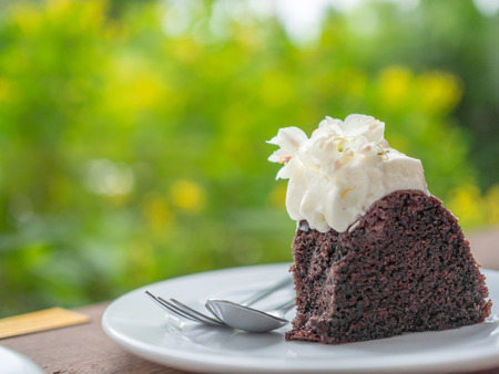 A piece of wild organic chocolate honey cake with beautiful vintage flowers topping and whipping cream on top in white ceramic plate with sillver spoon and fork on wooden table on green nature background.の写真素材