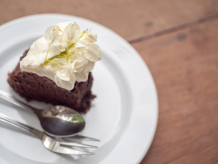 A piece of wild organic chocolate honey cake with beautiful vintage flowers topping and whipping cream on top in white ceramic plate with sillver spoon and fork on wooden table background.の写真素材