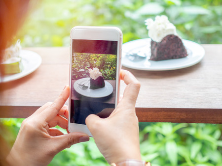 Hands shooting chocolate cake on white plate on wooden table from a smart phone camera on green garden background in cafe.の写真素材
