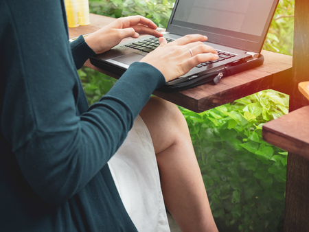 Woman wearing blue shirt and white skirt typingÂ laptopÂ computer on wooden counter bar and sittingÂ cross legsÂ near the window with garden view in the cafe.の写真素材