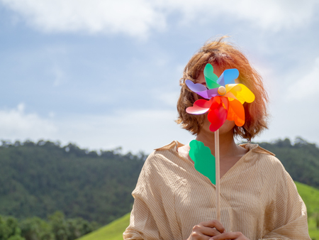 Woman wearing sunglasses and yellow relaxing shirt holding beautiful colorful pinwheel on green mountain landscape and blue sky background on sunny day with copy space.の写真素材
