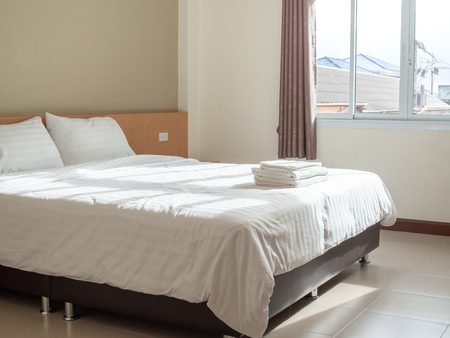 White clean bedroom in hotel, pillows and towels on the bed, curtains and window light from outside with houses view in countryside.の写真素材
