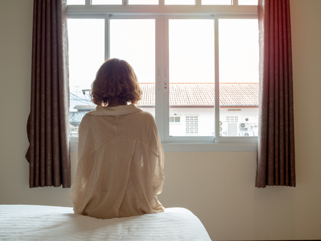Woman short hair sitting on the white bed in hotel room . Lonely woman looking out the window alone in white hotel room.の写真素材