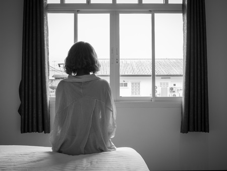 Woman short hair sitting on the white bed in hotel room . Lonely woman looking out the window alone in white hotel room, black and white style.