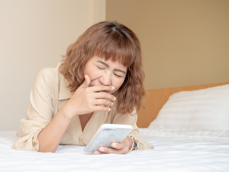 Beautiful asian woman short hair lying in white bed looking at smartphone and yawning.の写真素材