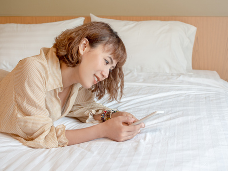 Beautiful happy smiling asian woman short hair lying in white bed using mobile phone with copy space.の写真素材