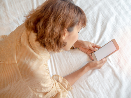 Beautiful happy smiling asian woman short hair lying in white bed using mobile phone top view.の写真素材