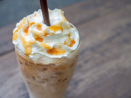 Close-up Iced Caramel Macchiato with Brown Straw in the tall glass. Cold coffee drink frappe (frappuccino) with whipped cream and caramel syrup and brown straw on wooden table background.の写真素材