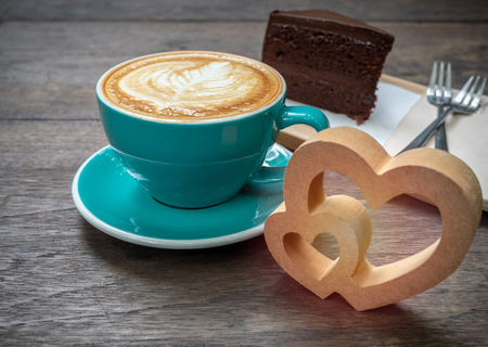 Coffee latte art in blue cup with a piece of chocolate cake, forks and wooden heart on wooden table background with space. Coffee lover concept.の写真素材