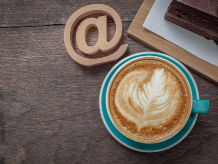 Flat lay coffee latte art in blue cup with wooden arroba symbol or email address icon and chocolate cake on wooden table background. Coffee lover and social network business concept on top view.の写真素材