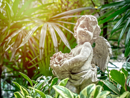 Old little vintage cupid statue holding the flowers in basket on green nature background with sunlight.の写真素材