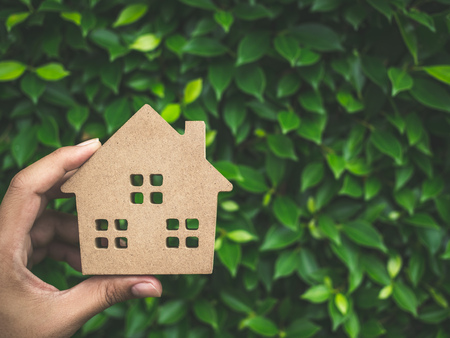 Hand holding wooden house on green leaves background with copy space. Home ecology concept.の写真素材