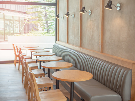 Row of wooden chairs, wooden round tables, built-in leather sofa and lamps on the concrete wall near the glass window wall in cafe with the light from outside on tree background.の写真素材