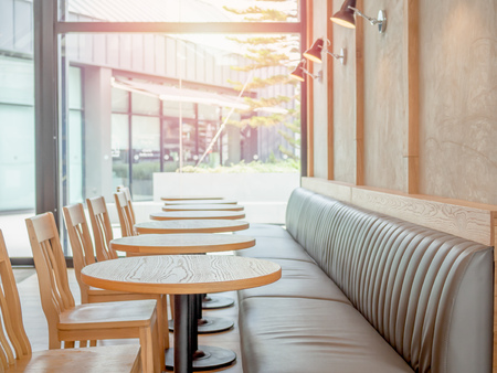 Row of wooden chairs, wooden round tables, built-in leather sofa and lamps on the concrete wall near the glass window wall in cafe with the light from outside on tree background.の写真素材