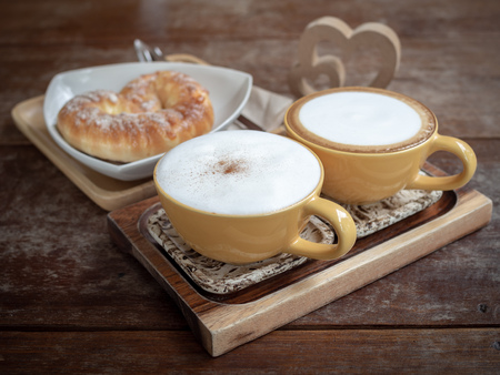Two hot coffee cups and sweet bread on white plate on wooden tray with wooden twin heart on wooden table background.の写真素材