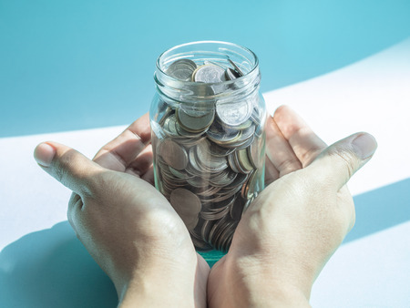 Coins in glass jar in hands isolated on blue and white background. Saving money and donation concept.の写真素材
