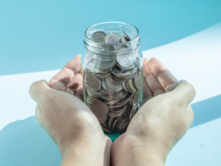 Coins in glass jar in hands isolated on blue and white background. Saving money and donation concept.の写真素材
