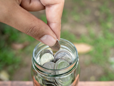 Close-up hand holding coin and put in glass jar on wooden table and on green tree background. Saving...