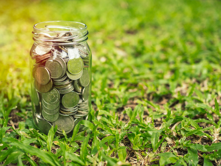 Coins in glass jar on green grass background with copy space. Saving money concept.の写真素材