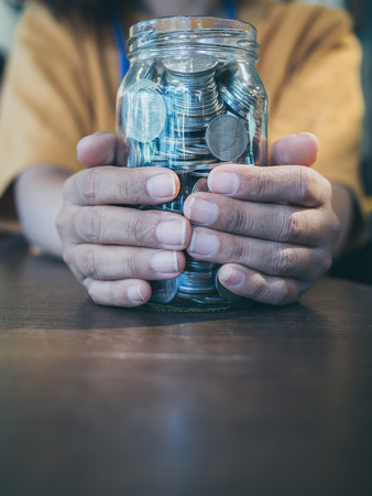 Coins in glass jar in hands of woman in yellow shirt on wooden table background vertical style. Saving money and donation concept.の写真素材