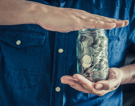 Man wearing blue shirt with hands holding coins in glass jar. Saving money and donation concept.の写真素材