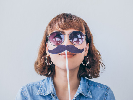 Beautiful asian woman short hair wearing blue jeans shirt and sunglasses smiling with fake mustache...