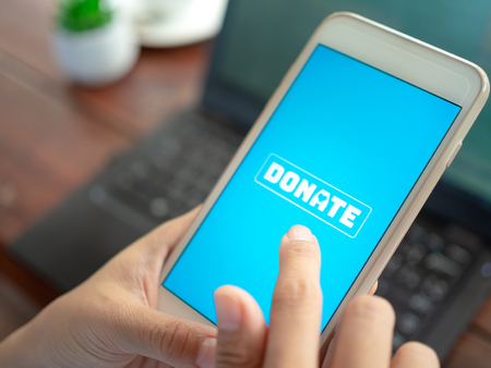 Close-up Finger pressing donate icon button on blue screen on white mobile phone with black laptop computer on wooden table. Donation online concept.の写真素材