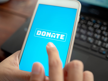 Close-up Finger pressing donate icon button on blue screen on white mobile phone with black laptop...