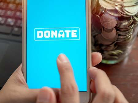 Close-up Finger pressing donate icon button on blue screen on white mobile phone with coins money in glass bottle with black laptop computer. Donation online concept.の写真素材