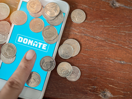 Finger pressing donate icon button on blue screen on white mobile phone with coins Thai baht money on wooden background with copy space. Donation online concept.の写真素材
