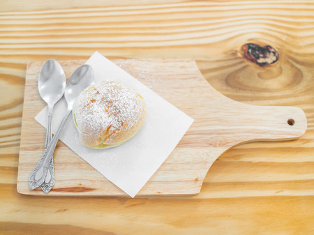 Delicious eclairs sprinkled with icing sugar and spoons on wooden cutting board on table top view.