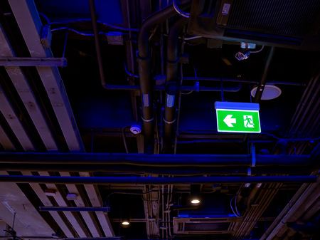 Illuminated green fire exit LED light sign on the ceiling in the dark with pipe line system inside the building.の写真素材