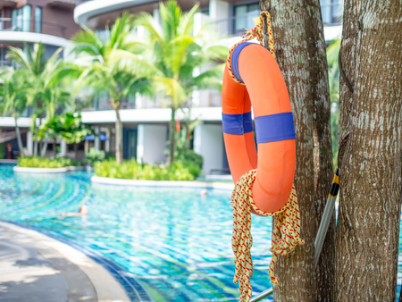 Orange lifebuoy with the rope hanging on the tree near the swimming pool and building.の写真素材