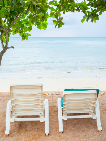 Two empty white plastic sunbeds with green pads and trees on sand beach on seascape background...