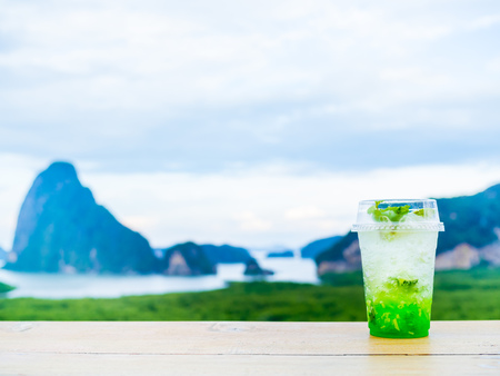 Iced lemon cocktail with lemon slice and peppermint leaf in transparent plastic glass on wooden table on seascape background with copy space. Sour green refreshing beverage.の写真素材
