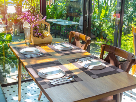 White plates, stainless steel spoons and forks on plate mats with bouquet purple flowers on wooden table. Dining table set in restaurant near glass window.の写真素材