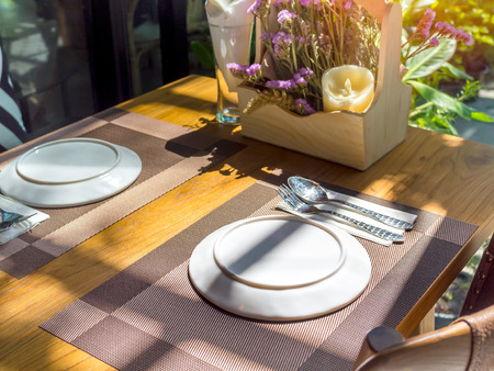 White plates, stainless steel spoons and forks on plate mats with bouquet purple flowers on wooden table. Dining table set in restaurant near glass window with sunshine.の写真素材