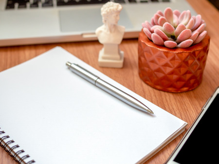 Close-up blank notebook with smartphone, silver pen, laptop computer, small david statue and succulent plant in copper pot on wooden table background vertical style. Office desk decoration.の写真素材