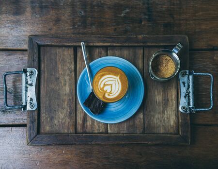 Flat lay Piccolo Latte coffee topping with flower art from milk in small glass with a piece of homemade brownies cake on blue ceramic plate and brown sugar in stainless container on wooden tray on wooden table.の写真素材
