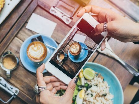 Woman's hands taking photo of coffee cup, Piccolo Latte, brownies with breakfast on wooden table by...