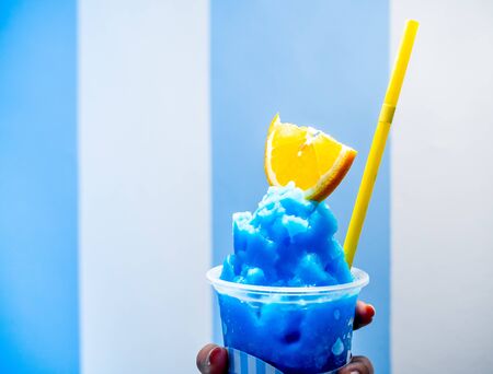 Close up hand holding fresh blue lemon smoothie with sliced yellow lime and yellow drinking straw in plastic cup on white and blue striped wall background.の写真素材