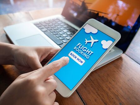 Flight booking online with smartphone. Finger touching on mobile phone for booking air flight online airline ticket with laptop computer on wooden table.の写真素材
