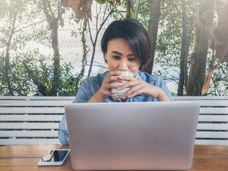 Beautiful happy asian woman short hair wearing blue jeans shirt drinking white cup of hot coffee and looking on laptop computer and her smartphone put on wooden table in cafe with green background.の写真素材