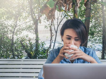 Beautiful happy asian woman short hair wearing blue jeans shirt drinking white cup of hot coffee and looking on laptop computer in cafe with green background.の写真素材