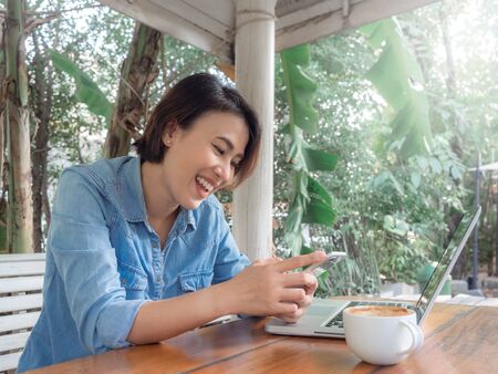 Beautiful laughing asian woman using smartphone with laptop computer and white hot coffee cup on wooden table in cafe.の写真素材