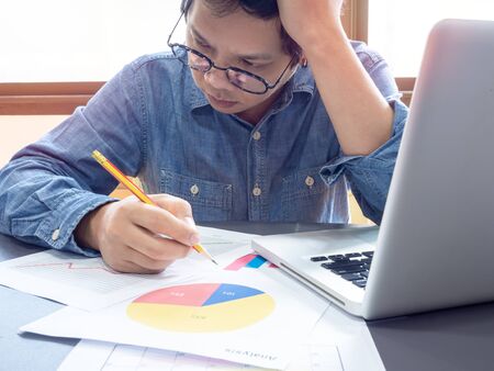 Asian businessman wearing blue shirt and glasses holding pencil and thinking with working with laptop computer on table near the window in office.の写真素材