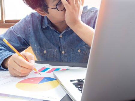 Asian businessman wearing blue shirt and glasses holding pencil and feel disappointed with working with laptop computer on table near the window in office.の写真素材
