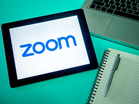 Bangkok, Thailand - April 17, 2020: "Zoom" logo on white background on tablet with notebook, pen and computer on blue background. "Zoom" is meetings application, video conference and communicationsのeditorial素材
